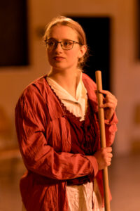 Junge Frau mit Brille und roter Jacke hält Stab in beiden Händen vor unscharfem Hintergrund