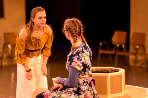 Zwei junge Frauen in einem Bühnenraum vor einem oktagonalen Holztisch, im Hintergrund leere Stühle.