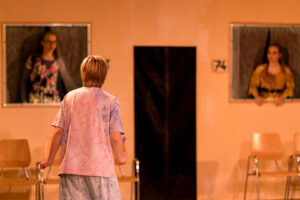 Junge von hinten vor orangefarbener Wand mit zwei Mädchen in Fenstern und leeren Holzstühlen.