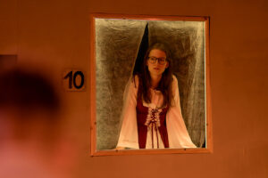 Junge Frau mit Brille in historischem Kleid steht in beleuchtetem Fensterrahmen mit Schild Nummer 10.