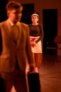 Junge Frau mit Schirmmütze und kariertem Schürze steht im Raum, unscharfe Person mit Aktenkoffer im Vordergrund.