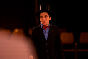 Junge in schwarzem Frack, roter Fliege und Zylinder vor dunklem Raum mit Stühlen.