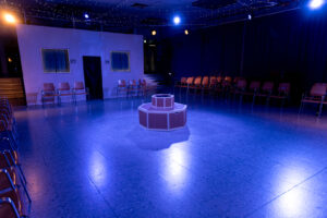 Theaterbühne mit Stühlen im Halbkreis, zweistufige Plattform, blaue Bühnenbeleuchtung, Wand mit Fenstern und Tür.