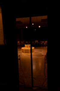 Dunkler Raum mit beleuchteter zweistufiger achteckiger Struktur und Stühlen im Halbkreis.