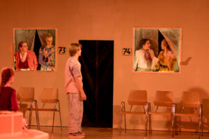 Bühne mit zwei Fensteröffnungen 73 und 74, Personen in Kostümen, Holzstühle im Vordergrund.