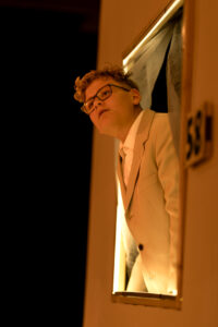 Junger Mann mit Brille trägt hellen Anzug und lehnt aus beleuchtetem Fenster in dunkler Umgebung.