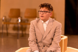 Junge mit Brille und beigem Anzug sitzt auf hölzernem Sitzmöbel vor Stühlen in Raum.