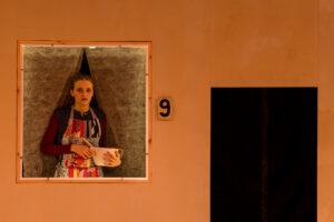 Frau in bunter Schürze steht mit Schüssel und Schneebesen in beleuchtetem Fenster Nr. 9.