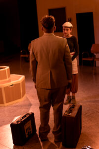 Mann im Anzug steht mit zwei Koffern vor einer jungen Frau mit weißer Kappe in warmem Licht.