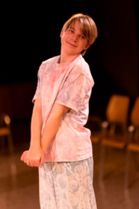 Junges Kind in Raum mit Stühlen trägt buntes T-Shirt und gemusterte Hose lächelnd zur Seite.