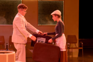 Zwei junge Personen übergeben sich in einem Raum jeweils einen Koffer vor Stühlen an der Wand.