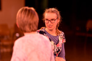 Zwei Frauen im Gespräch, eine mit Brille und floralem Kleid, die andere von hinten.