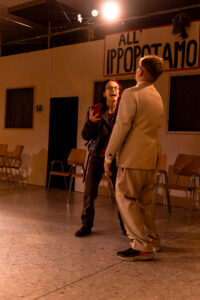 Zwei Personen im Gespräch vor einer Wand mit Schild ALL IPPOPOTAMO und mehreren Stühlen.