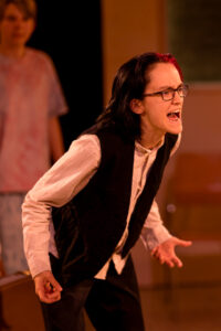 Person mit schwarzem Haar und Brille zeigt wütende Mimik und Gestik, Hintergrund unscharf.