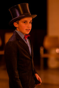 Junge mit schwarzem Zylinder, schwarzem Anzug, hellblauem Hemd und roter Fliege in warmem Licht.
