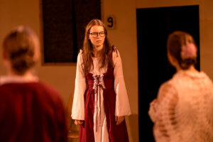 Junge Frau in historischem Kleid mit Brille steht einem Paar gegenüber in Kostümen.