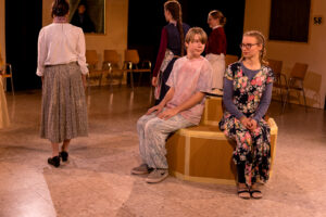 Jugendliche in Innenraum, einige sitzen auf runder Sitzgelegenheit, andere stehen, Stühle an der Wand.