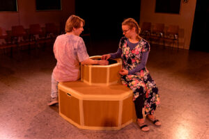 Zwei Personen sitzen auf sechseckiger Holzkonstruktion und halten Hände im Raum mit Stühlen.