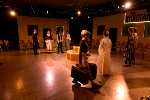 Theaterszene mit Schauspielern in historischen Kostümen, einer trägt zwei Koffer, eine Häkeldecke sichtbar.