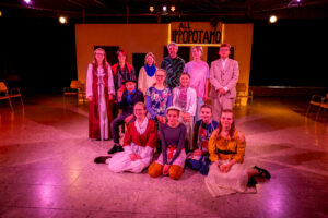 Jugendliche und Erwachsene in Theaterkostümen auf Bühne vor Schild All' Ippopotamo.