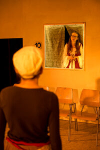 Person mit heller Kappe vor Spiegel, im Spiegel Frau im historischen Kleid, drei Stühle an oranger Wand mit Nummer 10.