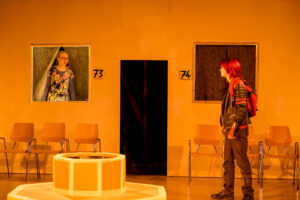 Theaterbühne mit zwei Personen, nummerierten Kabinen und Holzstühlen vor orangefarbener Wand.