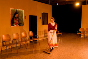 Frau in traditioneller Kleidung kehrt Boden, weitere Frau mit Schürze blickt aus Fenster, Holzstühle stehen links und rechts.
