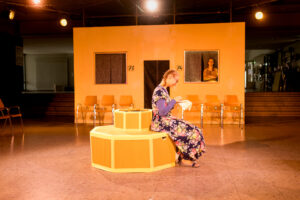 Frau in geblümtem Kleid sitzt auf gelbem sechseckigem Objekt und stickt in Innenraum.