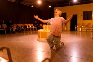 Junge kniet auf Bühnenboden in beleuchtetem Raum mit Stuhlreihen und hebt Arme.