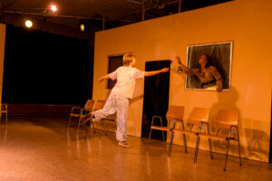 Zwei Personen in einem gelben Raum, eine reicht der anderen durch ein Fenster eine Blume.