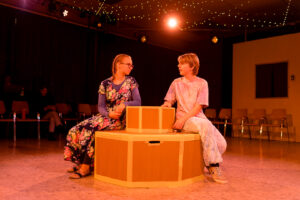 Zwei Kinder sitzen auf einem sechseckigen Podest aus Pappe in einem Raum mit Stuhlreihen.