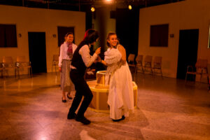 Drei Schauspieler proben eine Theaterszene auf einer Bühne mit nummerierten Türen und Stühlen.