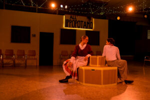 Zwei Personen in historischen Kostümen sitzen auf rundem Podest in Theatersaal mit Stühlen.