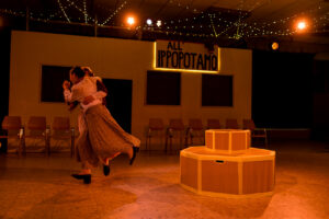 Zwei Personen tanzen auf einer Bühne mit Stühlen und beleuchtetem Schild im Hintergrund.